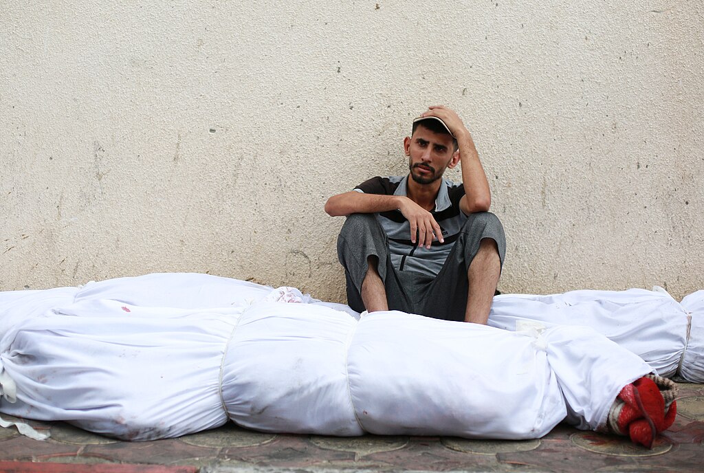 A Palestinian man sitting next to dead bodies wrapped in white sheets